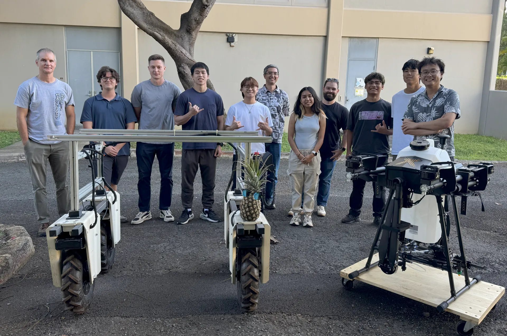 Ten guys and a young woman stand behind a 4-wheeled robot and a sprayer drone.